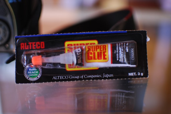 A tube of super glue in its packaging. The package is black with red and yellow accents and displays the brand name 'Alteco' prominently. The glue tube is visible through a clear section of the packaging, with a white and orange dispensing nozzle.