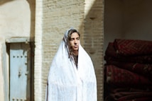 A woman stands wearing a light-colored shawl, with a thoughtful expression. She is positioned next to a stack of folded rugs and an old blue door with visible metal rivets. The background features a sunlit brick wall, creating a contrast between light and shadow.