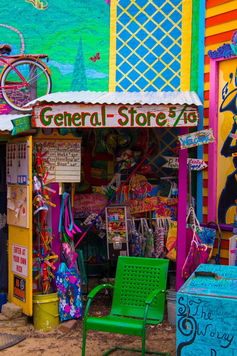 A vibrant and eclectic general store with colorful murals and signage. The storefront displays a mix of playful decorations, including a bicycle mounted on the wall and a variety of whimsical, hand-painted signs. A green perforated chair is in the foreground, and various merchandise is visible, such as bags and clothing. The overall scene is filled with bold colors and artistic design.