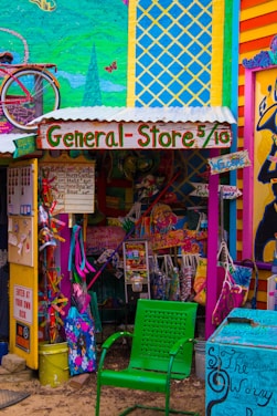 A vibrant and eclectic general store with colorful murals and signage. The storefront displays a mix of playful decorations, including a bicycle mounted on the wall and a variety of whimsical, hand-painted signs. A green perforated chair is in the foreground, and various merchandise is visible, such as bags and clothing. The overall scene is filled with bold colors and artistic design.