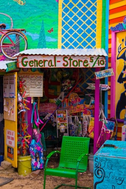 A vibrant and eclectic general store with colorful murals and signage. The storefront displays a mix of playful decorations, including a bicycle mounted on the wall and a variety of whimsical, hand-painted signs. A green perforated chair is in the foreground, and various merchandise is visible, such as bags and clothing. The overall scene is filled with bold colors and artistic design.