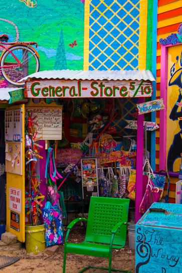 A vibrant and eclectic general store with colorful murals and signage. The storefront displays a mix of playful decorations, including a bicycle mounted on the wall and a variety of whimsical, hand-painted signs. A green perforated chair is in the foreground, and various merchandise is visible, such as bags and clothing. The overall scene is filled with bold colors and artistic design.