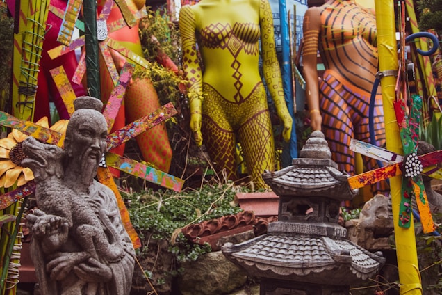 A collection of colorful mannequins in vibrant clothing patterns are surrounded by abstract art and garden elements. In the foreground, there is an ornate stone statue of a figure holding an animal beside a traditional Japanese lantern. The scene is lively and eclectic, combining natural greenery with vivid artificial elements.