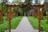 A garden with a series of wooden trellises lines a symmetrical stone pathway. The path leads to a statue in the distance surrounded by lush greenery. The scene is framed by vibrant green grass and various trees, including one with pink blossoms.