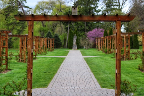 A garden with a series of wooden trellises lines a symmetrical stone pathway. The path leads to a statue in the distance surrounded by lush greenery. The scene is framed by vibrant green grass and various trees, including one with pink blossoms.