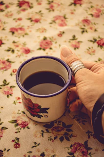 Close-up of a woman’s hand with delicate rings holding a lavender-hued coffee cup against a light beige background