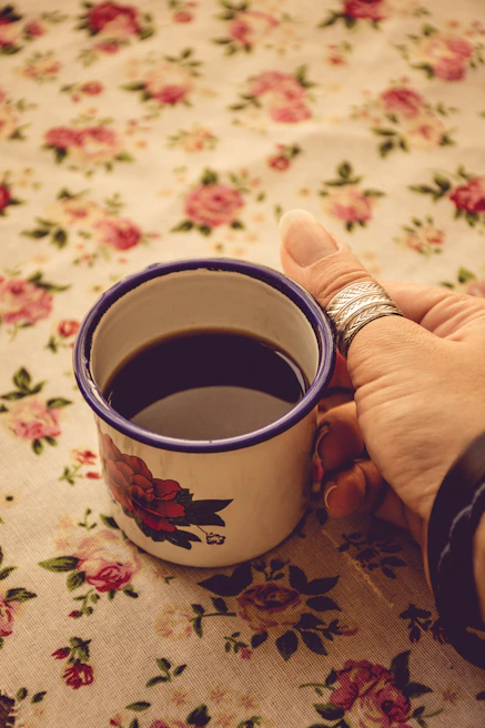 Close-up of a woman’s hand with delicate rings holding a lavender-hued coffee cup against a light beige background