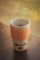 A reusable coffee cup with an orange textured grip sleeve sits on a wooden table. The cup has a simple design with a logo and arrow graphic. In the background, a blurred view of greenery can be seen through a window.