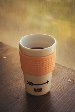 A reusable coffee cup with an orange textured grip sleeve sits on a wooden table. The cup has a simple design with a logo and arrow graphic. In the background, a blurred view of greenery can be seen through a window.