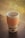 A reusable coffee cup with an orange textured grip sleeve sits on a wooden table. The cup has a simple design with a logo and arrow graphic. In the background, a blurred view of greenery can be seen through a window.
