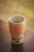 A reusable coffee cup with an orange textured grip sleeve sits on a wooden table. The cup has a simple design with a logo and arrow graphic. In the background, a blurred view of greenery can be seen through a window.