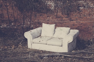 An old, worn-out white couch sits outdoors against a brick wall, surrounded by dry grass and twigs. The couch appears neglected, with visible stains and discoloration. Leafless branches and twigs create a stark contrast with the muted tones of the sofa and background.