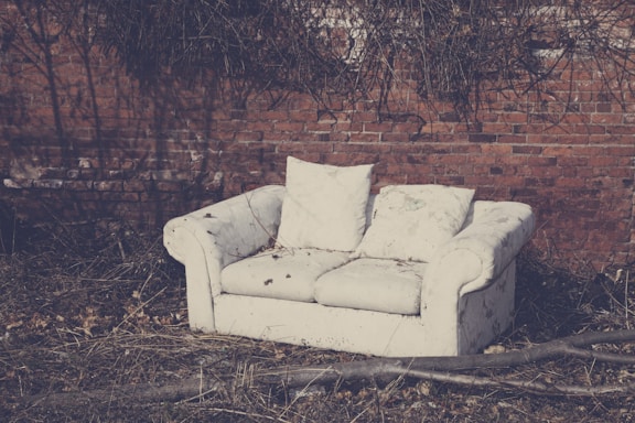 An old, worn-out white couch sits outdoors against a brick wall, surrounded by dry grass and twigs. The couch appears neglected, with visible stains and discoloration. Leafless branches and twigs create a stark contrast with the muted tones of the sofa and background.