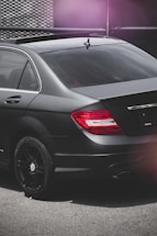 A close-up view of the rear section of a sleek black car parked on a concrete surface. The car features tinted windows and dark alloy wheels. The tail lights are illuminated in red, and the image has a moody lighting with a slight pink lens flare in the corner.