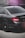 A close-up view of the rear section of a sleek black car parked on a concrete surface. The car features tinted windows and dark alloy wheels. The tail lights are illuminated in red, and the image has a moody lighting with a slight pink lens flare in the corner.