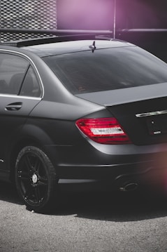 A close-up view of the rear section of a sleek black car parked on a concrete surface. The car features tinted windows and dark alloy wheels. The tail lights are illuminated in red, and the image has a moody lighting with a slight pink lens flare in the corner.