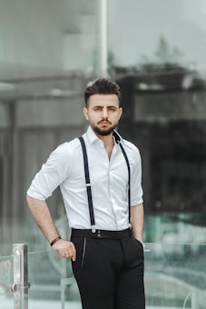 A man with a neatly styled beard and hair stands confidently, wearing a white shirt and black pants held up by suspenders. He is leaning on a glass railing in a modern, urban environment.
