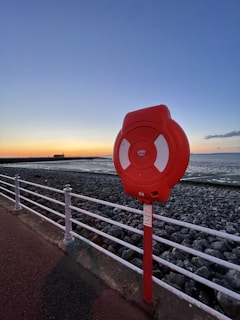 A red lifebuoy is mounted on a post along a seaside walkway. The background features a rocky shoreline with calm waters and a sunset sky painted in shades of blue and orange. A long white railing separates the path from the rocks.