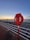 A red lifebuoy is mounted on a post along a seaside walkway. The background features a rocky shoreline with calm waters and a sunset sky painted in shades of blue and orange. A long white railing separates the path from the rocks.