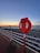 A red lifebuoy is mounted on a post along a seaside walkway. The background features a rocky shoreline with calm waters and a sunset sky painted in shades of blue and orange. A long white railing separates the path from the rocks.