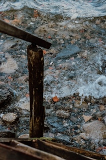 Weathered wooden beams stand near a rocky shoreline with water gently splashing against the stones. The scene captures the contrast between the rough textures of the aged wood and the smooth, fluid motion of the water.