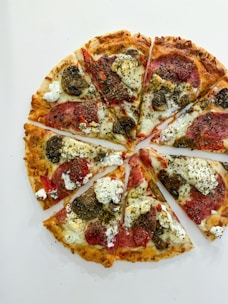 A pizza cut into eight slices is topped with ingredients including pepperoni, dollops of white cheese, mushrooms, and sprinkled herbs. The crust appears golden and crispy.