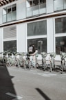 A person wearing a backpack stands next to a row of rental bicycles parked in front of a large building with numerous windows. The bicycles are mostly green, lined up in an organized manner, and situated in an urban setting. A shadow is cast across the pavement, suggesting sunlight overhead.