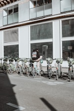 A person wearing a backpack stands next to a row of rental bicycles parked in front of a large building with numerous windows. The bicycles are mostly green, lined up in an organized manner, and situated in an urban setting. A shadow is cast across the pavement, suggesting sunlight overhead.