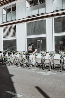 A person wearing a backpack stands next to a row of rental bicycles parked in front of a large building with numerous windows. The bicycles are mostly green, lined up in an organized manner, and situated in an urban setting. A shadow is cast across the pavement, suggesting sunlight overhead.