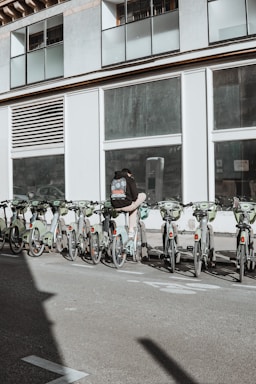 A person wearing a backpack stands next to a row of rental bicycles parked in front of a large building with numerous windows. The bicycles are mostly green, lined up in an organized manner, and situated in an urban setting. A shadow is cast across the pavement, suggesting sunlight overhead.