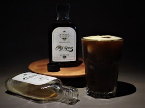 A bottle labeled 'Acacia Coffee Cold Brew' stands on a wooden surface next to a glass filled with dark cold brew coffee. An empty bottle with a loose cap lies on the surface, with a shadow cast behind it against a dark background.