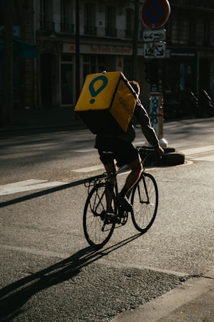 A cyclist rides on a city street carrying a large, yellow delivery backpack with a logo. The scene captures the early morning or late afternoon light, casting long shadows on the road. Urban buildings and a few parked vehicles are visible in the background.