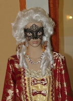 A mannequin dressed in a lavish historical costume featuring a red and gold patterned fabric with lace and bows. The figure has an ornate black lace mask covering part of the face and wears a white, curly wig styled in a vintage fashion. The mannequin is also adorned with a colorful beaded necklace.