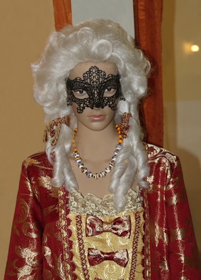 A mannequin dressed in a lavish historical costume featuring a red and gold patterned fabric with lace and bows. The figure has an ornate black lace mask covering part of the face and wears a white, curly wig styled in a vintage fashion. The mannequin is also adorned with a colorful beaded necklace.