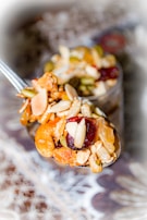 A close-up view of a spoon filled with assorted dried fruits and nuts, including almonds, cashews, pumpkin seeds, and dried cherries. The spoon is in focus with a blurred decorative background.