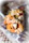 A close-up view of a spoon filled with assorted dried fruits and nuts, including almonds, cashews, pumpkin seeds, and dried cherries. The spoon is in focus with a blurred decorative background.