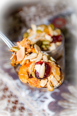 A close-up view of a spoon filled with assorted dried fruits and nuts, including almonds, cashews, pumpkin seeds, and dried cherries. The spoon is in focus with a blurred decorative background.