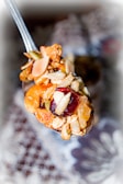 A close-up of a spoonful of mixed nuts, dried fruits, and grains. The mix includes almonds, cashews, raisins, and seeds. The background is blurred, drawing focus to the spoon's contents.