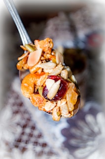 A close-up of a spoonful of mixed nuts, dried fruits, and grains. The mix includes almonds, cashews, raisins, and seeds. The background is blurred, drawing focus to the spoon's contents.