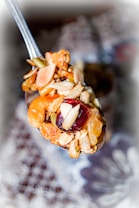 A close-up of a spoonful of mixed nuts, dried fruits, and grains. The mix includes almonds, cashews, raisins, and seeds. The background is blurred, drawing focus to the spoon's contents.