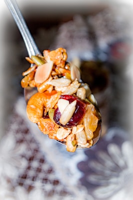 A close-up of a spoonful of mixed nuts, dried fruits, and grains. The mix includes almonds, cashews, raisins, and seeds. The background is blurred, drawing focus to the spoon's contents.