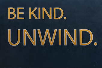 Bold yellow text on a dark background reads 'BE KIND. UNWIND.' The words are evenly spaced and capitalized, conveying a message of encouragement and relaxation.