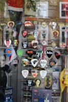 A selection of guitar picks with various rock band logos and designs are displayed against a window. Bands represented include The Beatles, AC/DC, Metallica, Nirvana, The Rolling Stones, and more. There are also logos from popular culture references like Superman and Doctor Who. The reflection of a street scene is visible in the glass along with some blurred brick buildings in the background.