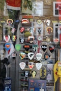A selection of guitar picks with various rock band logos and designs are displayed against a window. Bands represented include The Beatles, AC/DC, Metallica, Nirvana, The Rolling Stones, and more. There are also logos from popular culture references like Superman and Doctor Who. The reflection of a street scene is visible in the glass along with some blurred brick buildings in the background.