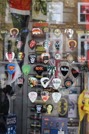 A selection of guitar picks with various rock band logos and designs are displayed against a window. Bands represented include The Beatles, AC/DC, Metallica, Nirvana, The Rolling Stones, and more. There are also logos from popular culture references like Superman and Doctor Who. The reflection of a street scene is visible in the glass along with some blurred brick buildings in the background.