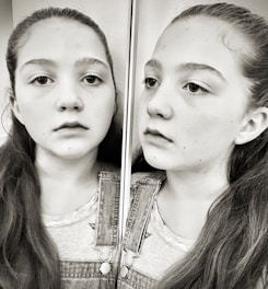 A black and white image features a young girl with long hair wearing a denim overall. She is looking at her reflection in a mirror, capturing a thoughtful or introspective expression.