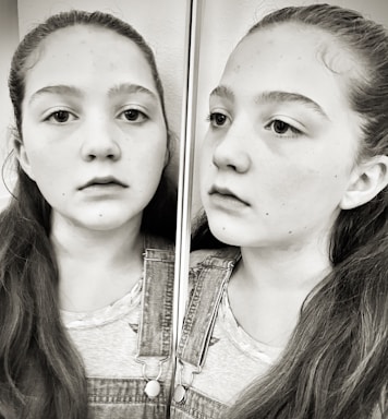 A black and white image features a young girl with long hair wearing a denim overall. She is looking at her reflection in a mirror, capturing a thoughtful or introspective expression.