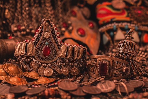 A collection of ornate, intricately designed ethnic jewelry featuring elaborate patterns, gemstones, and metalwork. The jewelry includes pieces with large red stones, detailed engravings, and a mix of gold and silver tones, all set against a richly textured background.