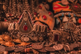 A collection of ornate, intricately designed ethnic jewelry featuring elaborate patterns, gemstones, and metalwork. The jewelry includes pieces with large red stones, detailed engravings, and a mix of gold and silver tones, all set against a richly textured background.