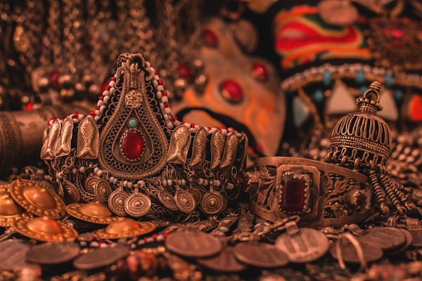 A collection of ornate, intricately designed ethnic jewelry featuring elaborate patterns, gemstones, and metalwork. The jewelry includes pieces with large red stones, detailed engravings, and a mix of gold and silver tones, all set against a richly textured background.